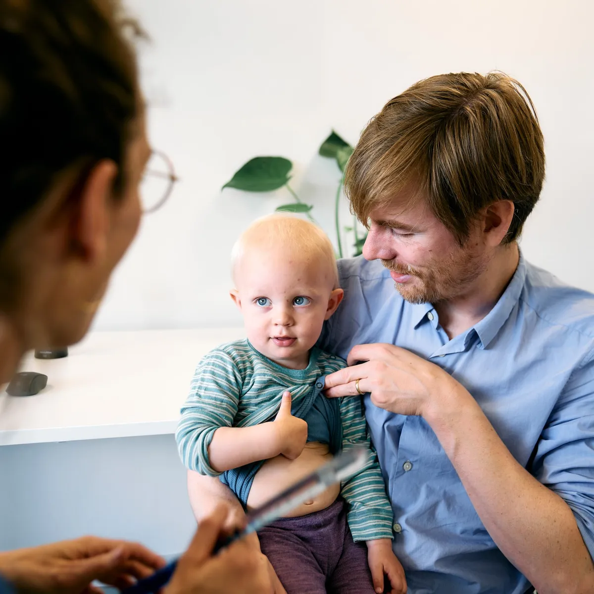 En far sidder med sit lille barn på skødet under en lægekonsultation.
